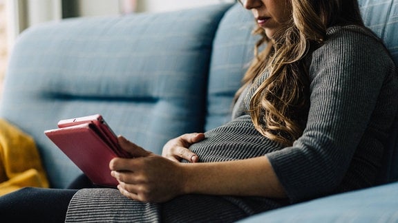 Abgeschnittene, nicht erkennbare Frau, die ein Baby erwartet, sitzt auf einem karierten Sofa und surft auf einem Tablet zu Hause.
