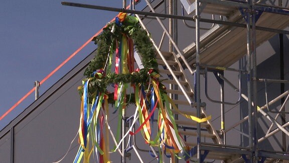 Eine Richtkrone aus Nadelzweigen mit bunten Bändern geschmückt an einem Baugerüst, dahinter ein Gebäude im Rohbau